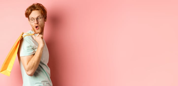 Carefree young man with red hair and glasses, walking with shopping bag, looking behind his shoulder with amazed expression, standing over pink background photo