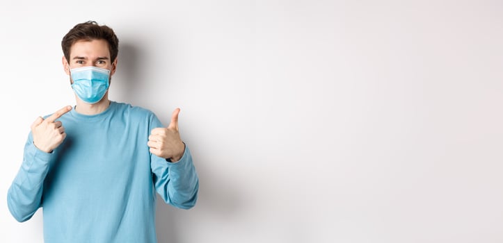 Coronavirus, health and quarantine concept. Smiling man pointing at his medical mask and showing thumb up, standing on white background photo