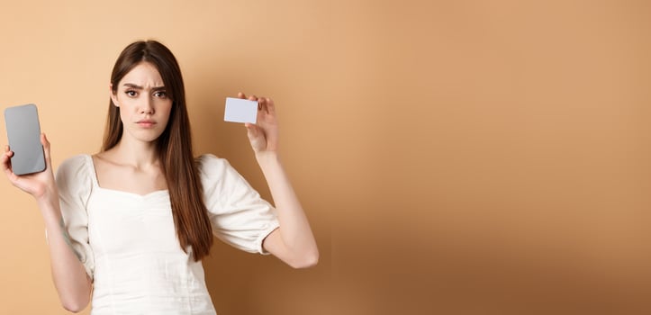 Sad and disappointed girl showing empty smartphone screen and plastic credit card, complaining and frowning, standing on beige background photo