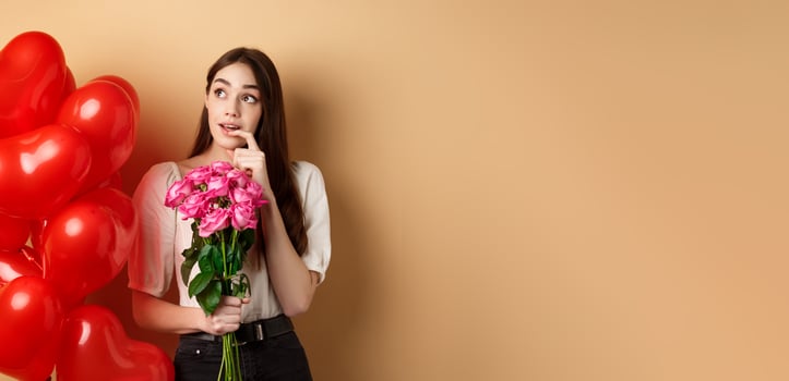Dreamy young woman holding bouquet of roses and thinking about secret admirer on Valentines day, looking at upper left corner and biting finger, standing near red romantic balloons photo