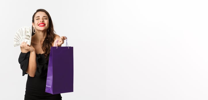 Satisfied and happy woman enjoying shopping, holding bag and money, smiling pleased, standing over white background photo