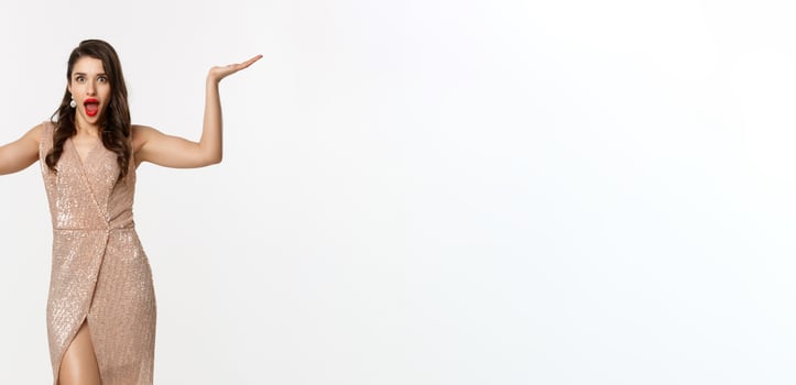 Party and celebration concept. Full-length of beautiful woman in elegant dress, standing near Christmas gifts and looking surprised, standing over white background photo