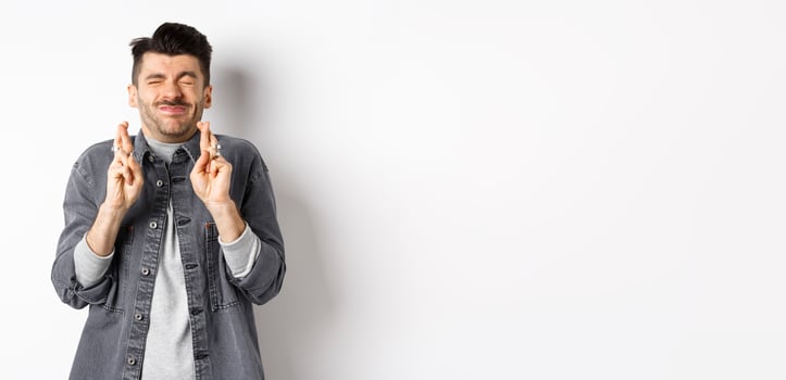 Hopeful young man standing nervous with closed eyes and fingers crossed, making wish, begging god for dream come true, waiting news, standing on white background photo