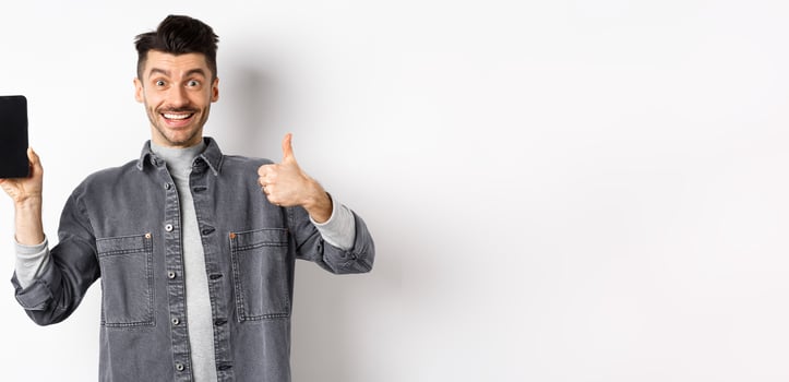 Cheerful stylish man showing empty smartphone screen and thumbs-up, concept of online shopping and e-commerce photo