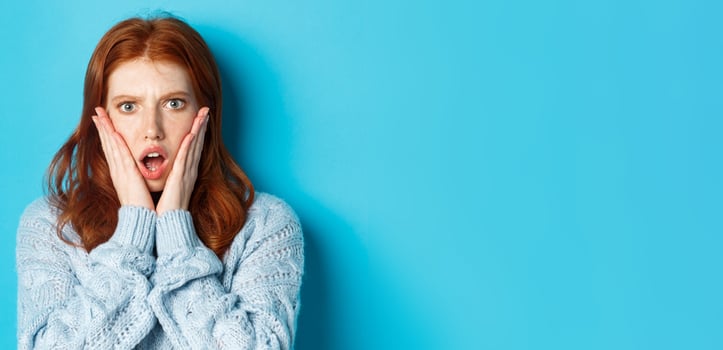 Close-up of shocked redhead girl staring at something displeasing, holding hands on face and gasping, standing over blue background photo