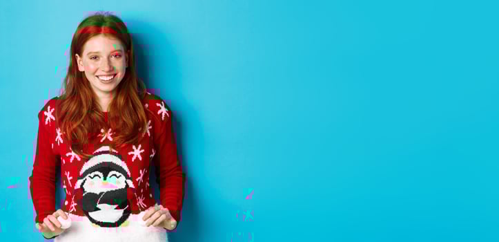 Winter holidays and Christmas Eve concept. Cute redhead girl wearing adorable sweater and smiling, celebrating New Year, standing over blue background photo