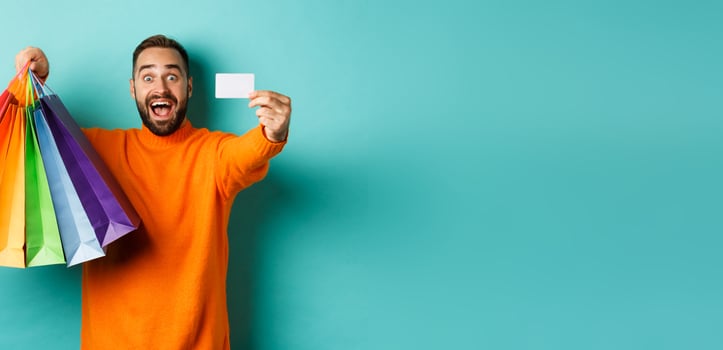Happy aduly man showing credit card and shopping bags, standing against turquoise background photo