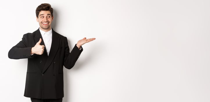 Handsome businessman in black suit, showing thumb-up and holding your product in hand over white copy space, standing against white background photo