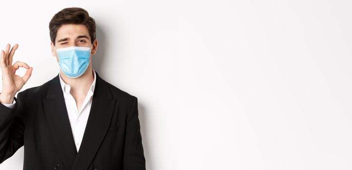 Concept of covid-19, business and social distancing. Close-up of good-looking businessman in trendy suit and medical mask, showing okay sign and winking, standing against white background photo