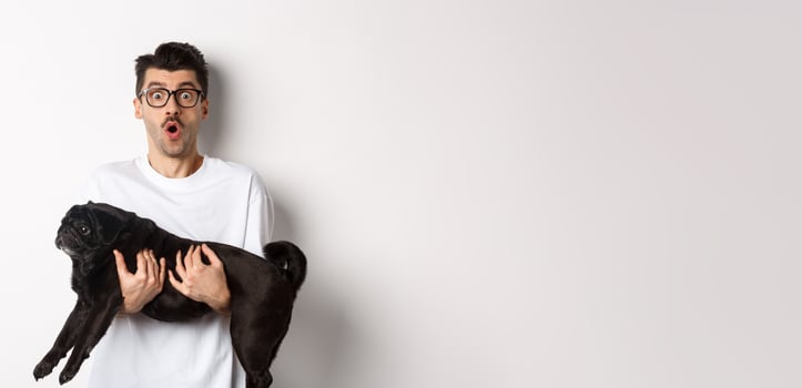Surprised young man in glasses holding cute black pug, dog owner staring at camera with impressed face, saying wow, standing over white background photo