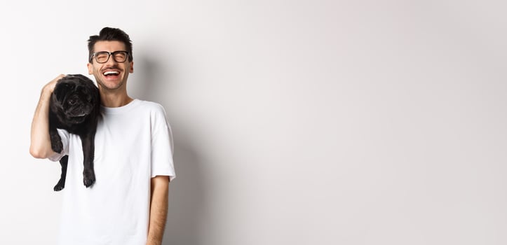 Happy young man laughing carefree, holding cute black dog, pug breed, on shoulder and having fun, standing over white background photo