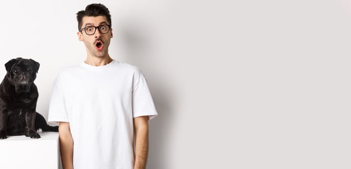 Image of dog owner and cute black pug looking at camera surprised and amazed, standing over white background photo
