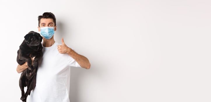 Covid-19, animals and quarantine concept. Young man in medical mask holding cute black pug dog, showing thumb up, like and approve, standing over white background photo