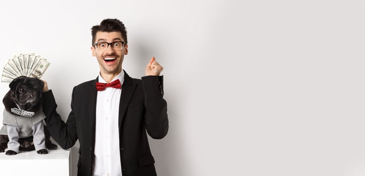Happy man winning money, wearing costume and showing dollars near his cute black dog in suit, standing over white background photo