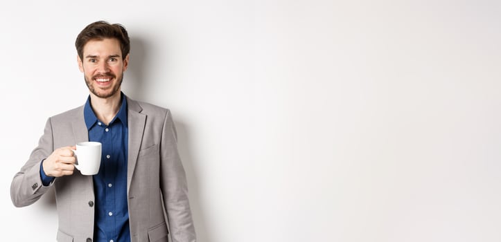 Handsome businessman in suit drinking coffee or tea from office mug, smiling enthusiastic at camera, standing against white background photo