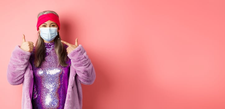 Covid-19, social distancing and fashion concept. Smiling asian senior woman in fashionable clothes and respirator, pointing at face mask and show thumb-up, pink background photo