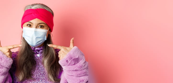 Covid-19, virus and fashion concept. Fashionable asian senior woman in trendy outfit pointing fingers at medical mask, follow social distancing rules, pink background photo