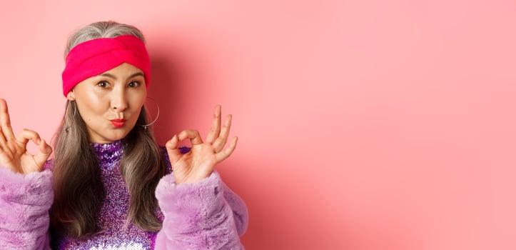 Fashion. Coquettish asian senior woman in trendy headband and faux fur coat, showing okay signs and pucker lips in kiss, praising something perfect, pink background photo