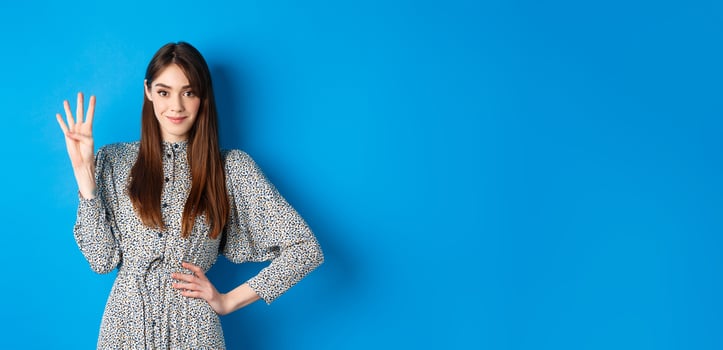Young beautiful girl in dress with natural long hair, showing number four with fingers and smiling, standing against blue background photo