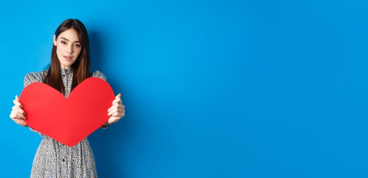 Valentines day and relationship concept. Tender young woman in dress stretch out hand and giving big red heart to you, making confession, standing romantic on blue background photo