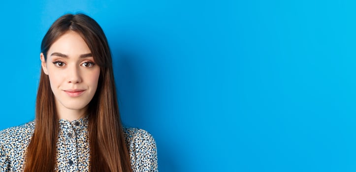 Hopeful candid woman with long hair and makeup, looking at camera with smile, standing against blue background photo