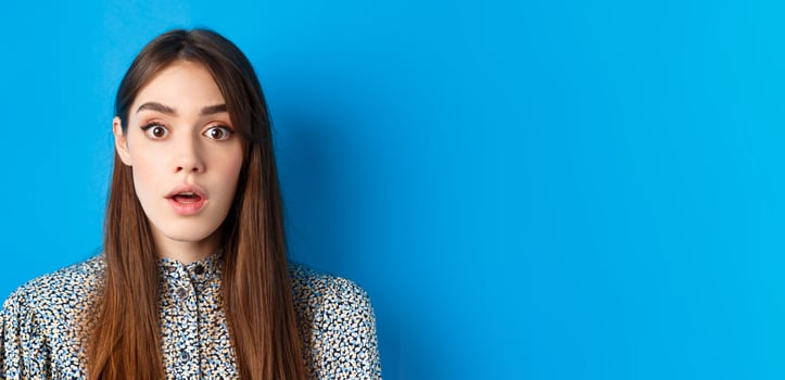 Close-up of excited pretty woman with long natural hair and makeup, drop jaw and gasping surprised, checking out awesome promo, hear amazing news, blue background photo