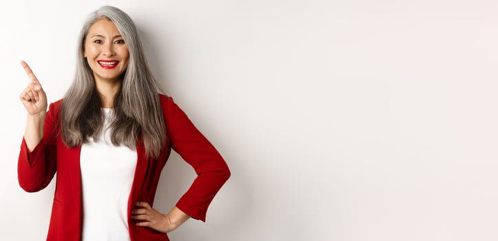 Happy senior office lady pointing finger at upper left corner, showing company logo and smiling at camera, standing over white background photo