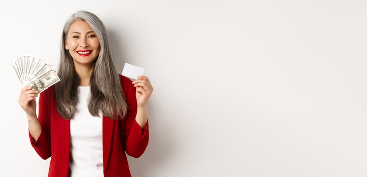 Successful asian senior businesswoman showing money in dollars and plastic card, smiling happy at camera, wearing red blazer and make-up photo