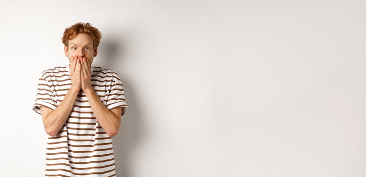 Surprised and excited man with curly red hair checking out special promo offer,staring fascinated and amazed, covering mouth with hands, standing over white background photo