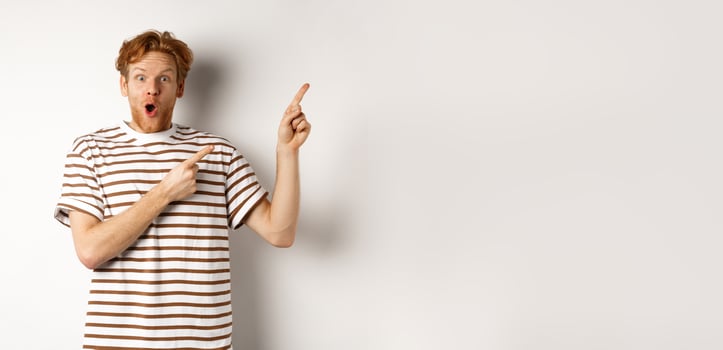 Excited man with curly red hair showing promo offer and looking amazed, pointing fingers at upper right corner logo, white background photo
