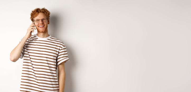 Hipster with red hair and glasses talking on mobile phone, smiling during conversation, white background photo