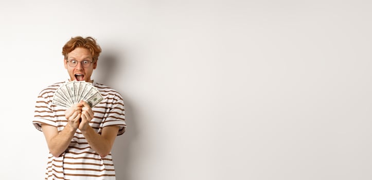 Lucky young man with red hair showing dollars, winning money and screaming of happiness, holding prize cash, standing over white background photo