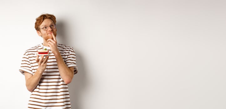 Holidays and celebration concept. Happy young man with red hair and glasses having birthday, holding cake with candle and thinking of b-day wish, standing over white background photo