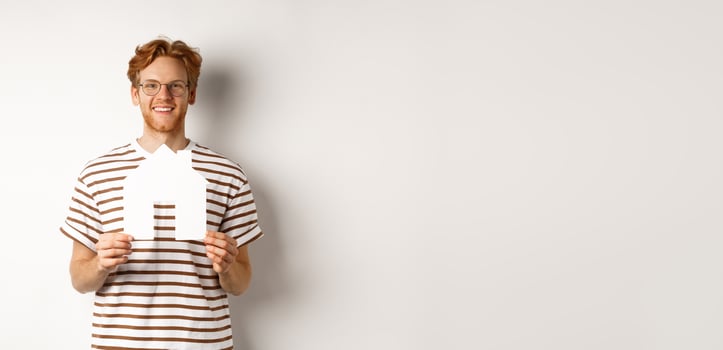 Real estate concept. Smiling redhead man in glasses holding cutout paper house and looking at camera, saving for buying property, standing over white background photo