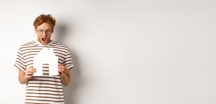 Amazed young man with red hair, staring at paper house cutout with excitement, standing over white background photo