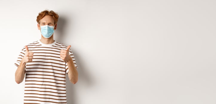 Covid-19, pandemic and social distancing concept. Young man with red hair wearing medical mask to prevent catching coronavirus, showing thumbs-up, white background photo