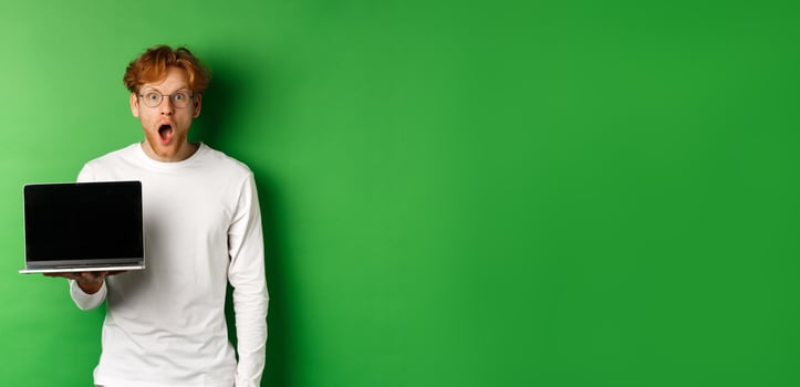 Impressed redhead guy in glasses drop jaw after seeing online promo, showing laptop screen and staring at camera with disbelief, standing over green background photo