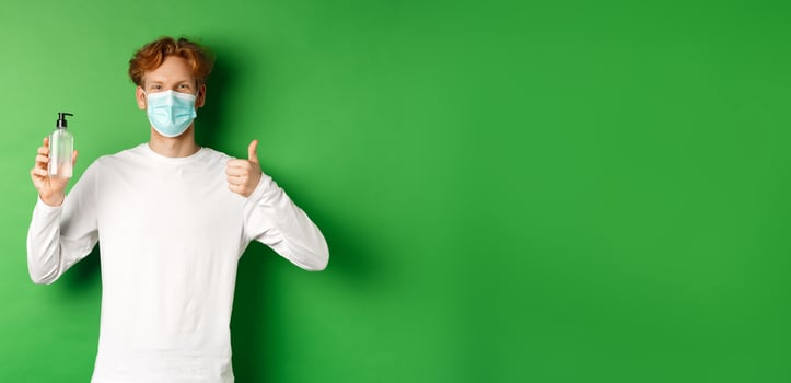 Covid-19, virus and social distancing concept. Smiling young man with red hair, wearing face mask for protection from corona, showing thumb-up and hand sanitizer, green background photo