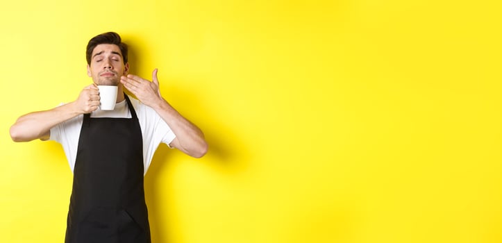 Barista enjoying smell of coffee in mug, standing pleased with eyes closed, wearing black apron photo