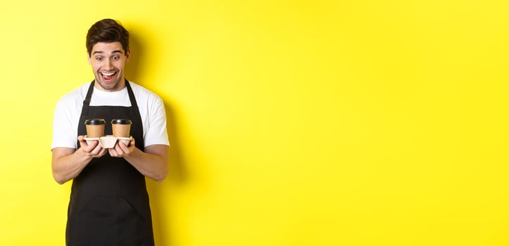 Waiter looking excited at two cups of takeaway coffee, wearing black apron, standing over yellow background photo