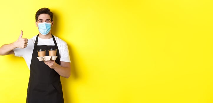 Concept of covid-19, cafe and social distancing. Young male barista in medical mask and black apron holding takeaway coffee cups, showing thumb up, yellow background photo
