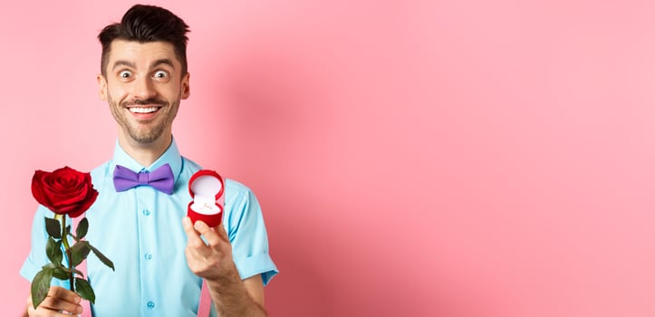 Valentines day. Smiling handsome man asking to marry him, showing engagement ring and red rose, standing romantic on pink background photo