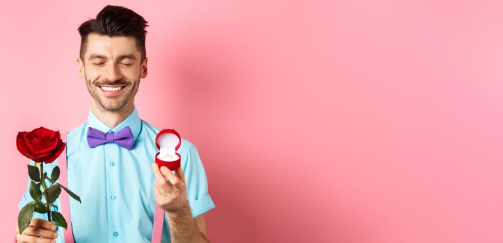 Valentines day. Cute boyfriend making wedding proposal, showing engagement ring in small box and red rose, express love, standing over pink background photo