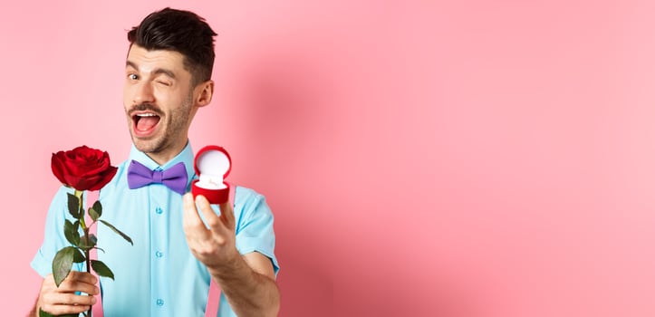 Valentines day. Funny guy making proposal, winking and saying marry me, showing engagement ring with red rose, standing over pink background photo
