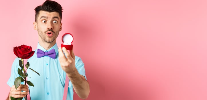 Valentines day. Romantic funny guy going to make wedding proposal, asking to marry him, holding red rose and looking at engagement ring with excitement, pink background photo