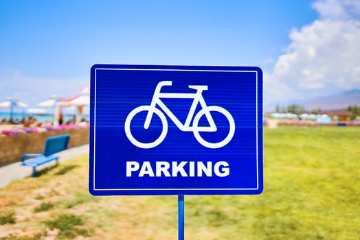 A vibrant blue traffic sign featuring a silhouette of a bicycle wheel parked against a sky backdrop with fluffy clouds. The font stands out against the natural landscape photo