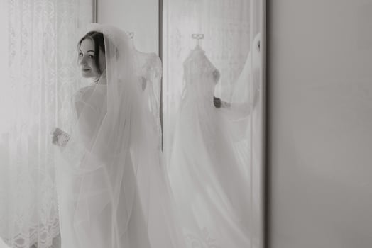 A woman in a wedding dress is standing in front of a mirror, adjusting her veil. The image has a nostalgic and romantic feel, as it captures a moment of preparation for a special occasion photo