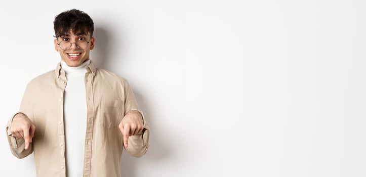 Portrait of happy young guy in glasses showing cool promo, pointing fingers down and smiling excited, standing on white background photo