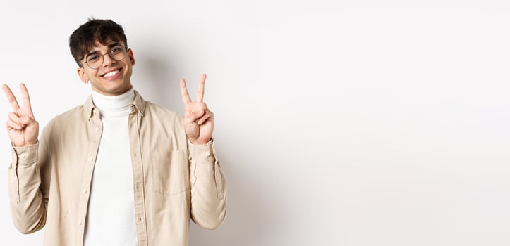Handsome hipster guy in glasses, smiling with white teeth, showing peace or victory gesture, standing upbeat on white background photo