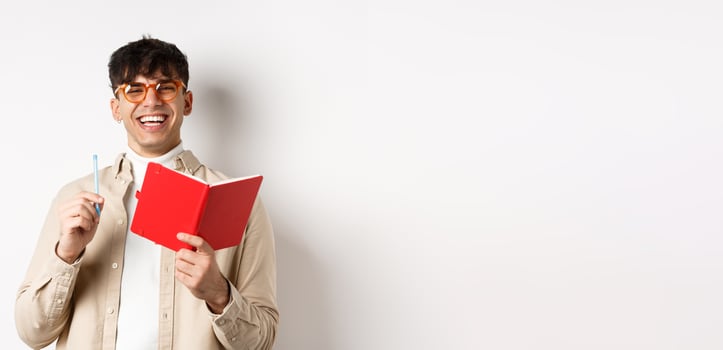 Cheerful young man in glasses laughing and taking notes, writing down in planner, holding pen and diary, standing on white background photo